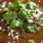 Thick, dark brown Mexican mole sauce in a clay bowl with chilies, chocolate, sesame seeds, and spices around it.
