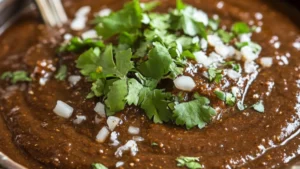 Thick, dark brown Mexican mole sauce in a clay bowl with chilies, chocolate, sesame seeds, and spices around it.